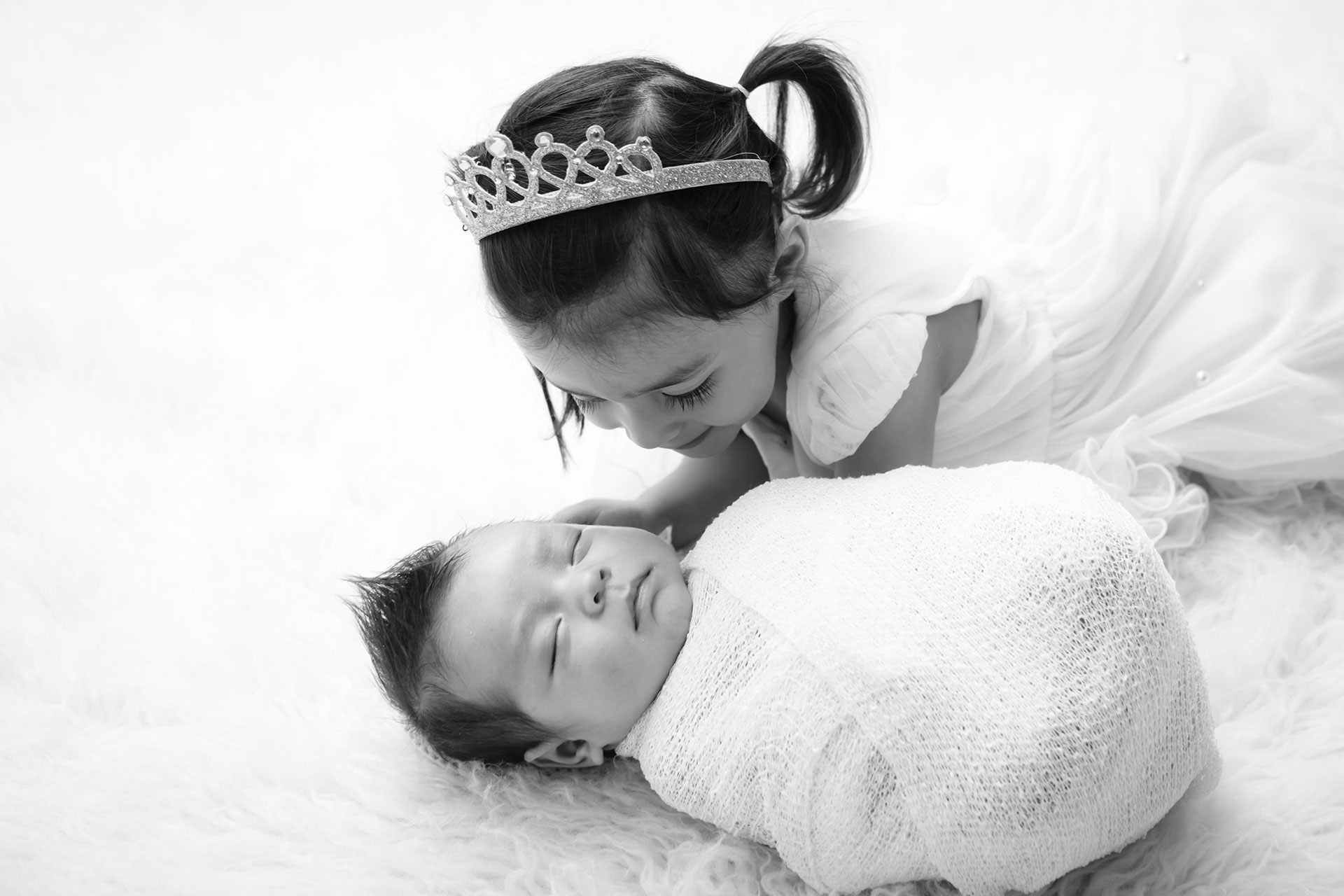 Older sister leaning close to her sleeping newborn sibling in a bright studio portrait